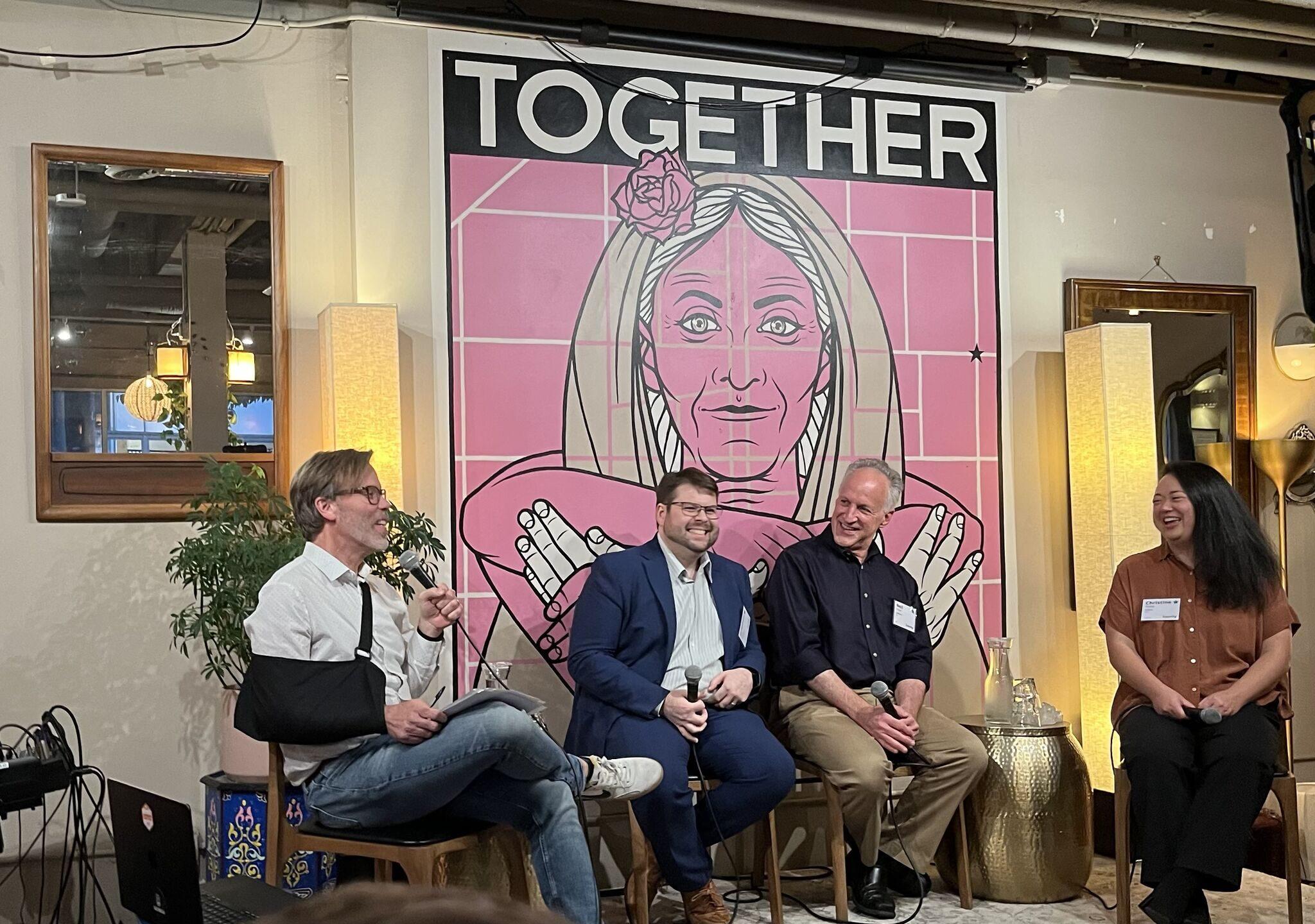 Christine Tsang, far right, laughing on stage with the moderator and two fellow panelists in front of a pink mural that reads “TOGETHER.”